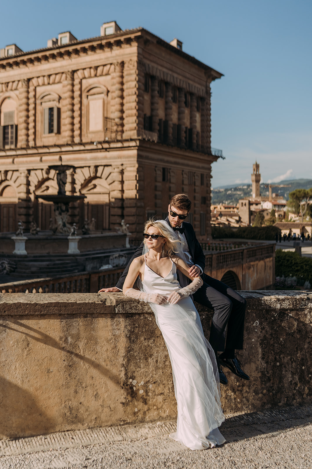 A bride in an elegant white satin gown leans against a stone terrace balustrade with her groom beside her, with the grand facade of Palazzo Pitti and the Florence skyline visible behind them.