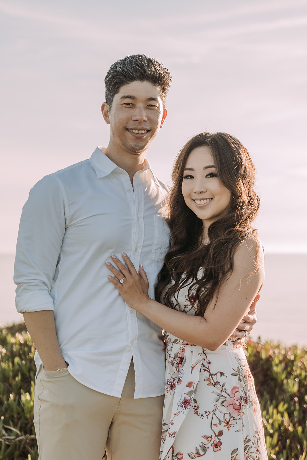 A couple smiles warmly at the camera in a close portrait, with coastal greenery softly blurred in the background. The woman wears a floral dress and leans into the man, who has his arm around her. An engagement ring is visible on her hand.