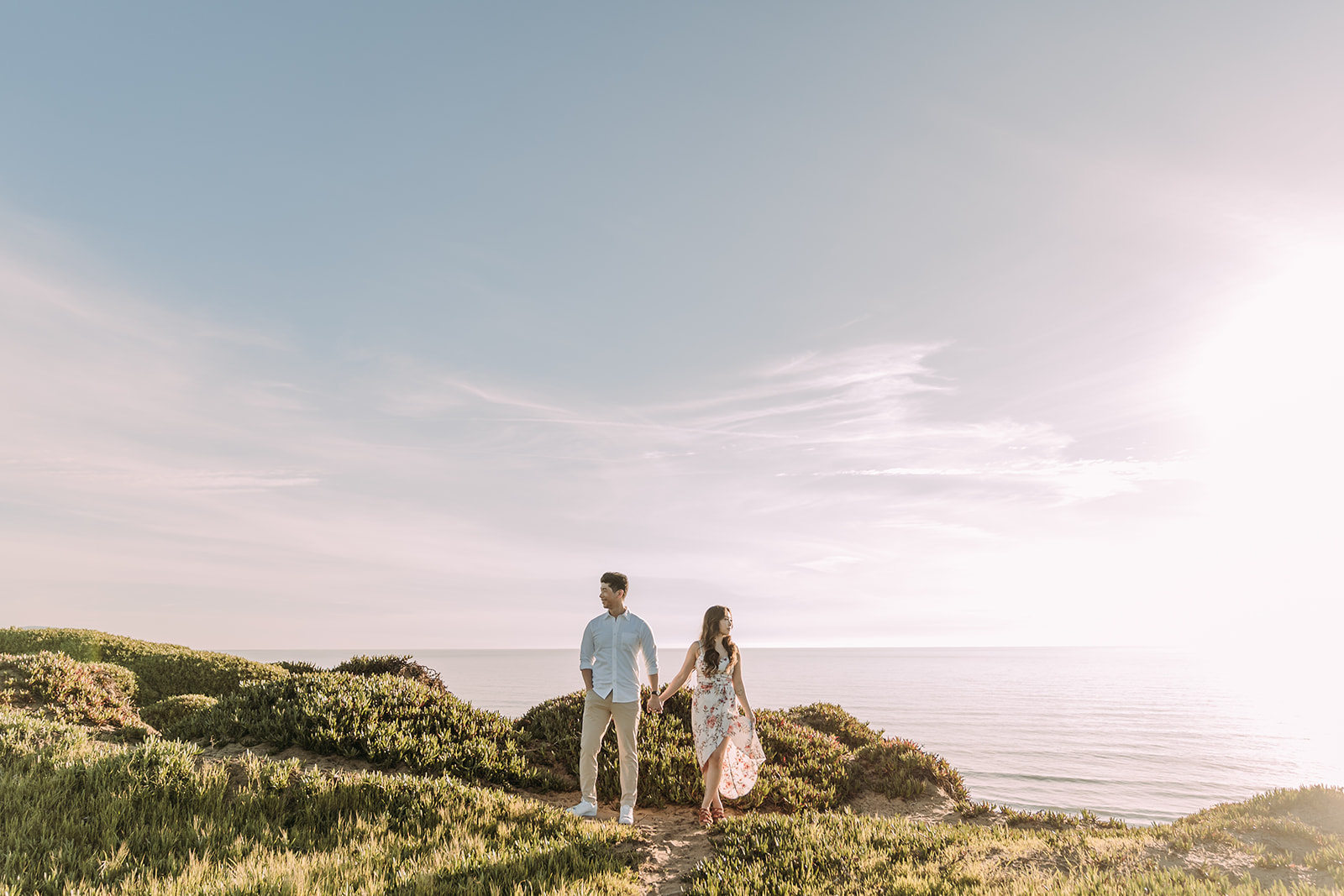 A couple holds hands and walks along a coastal cliffside trail at Fort Funston, surrounded by low green shrubs with a calm ocean and soft, hazy sky stretching behind them. The woman wears a floral dress and the man is in a light blue shirt and khakis.