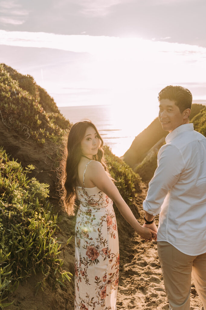 A couple holds hands and walks along a sandy coastal trail framed by lush greenery, with the sun setting dramatically behind them over the ocean. The woman looks back over her shoulder toward the camera.